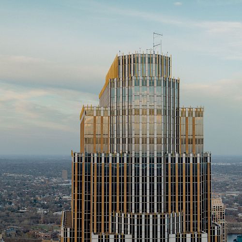 A tall building with a unique architectural design, featuring tiered gold accents and large windows, stands out against a cityscape background.