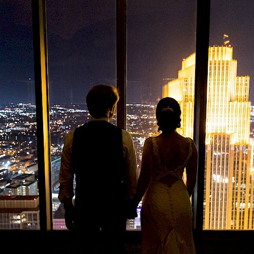 A couple stands hand-in-hand, gazing out a window at a brightly lit skyscraper and cityscape at night.