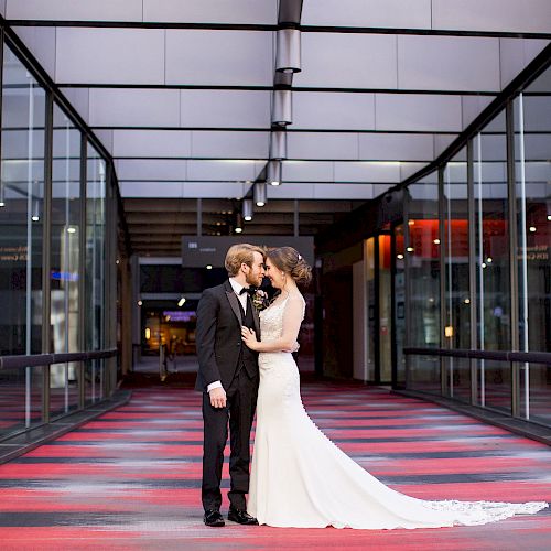 A bride and groom stand in an enclosed walkway, dressed in a wedding gown and tuxedo, holding each other closely, and sharing an intimate moment.