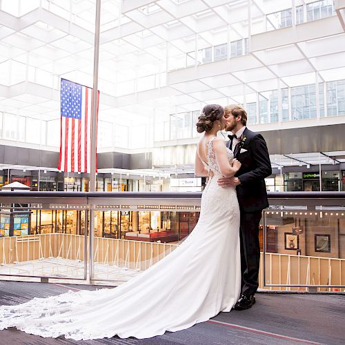 A couple in wedding attire share a moment near an indoor balcony with an American flag in the background, within a bright, modern building.