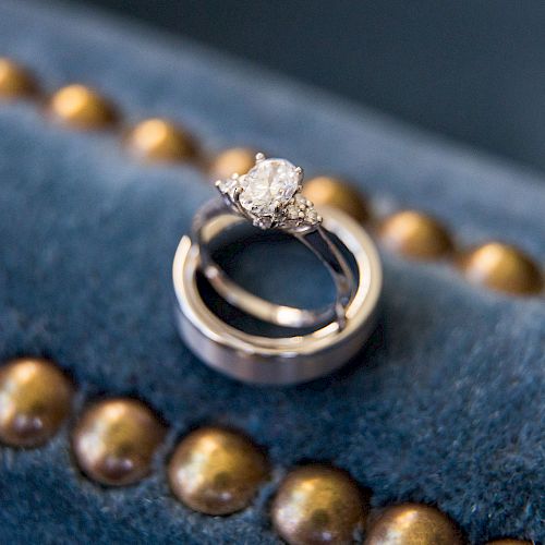 A pair of rings, including a diamond engagement ring, are placed on a blue cushion with golden accents.