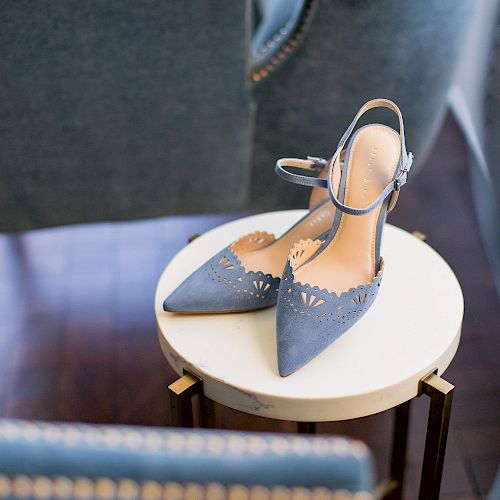 A pair of blue pointed-toe high heel shoes with cut-out details is placed on a small round table, surrounded by upholstered chairs.