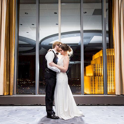 A couple dressed in formal attire, possibly at a wedding, embrace lovingly in front of a large window with city lights in the background.