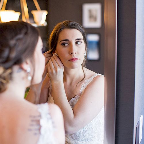 A bride is adjusting her earring in front of a mirror, her reflection clearly visible while she gets ready in a softly lit room.