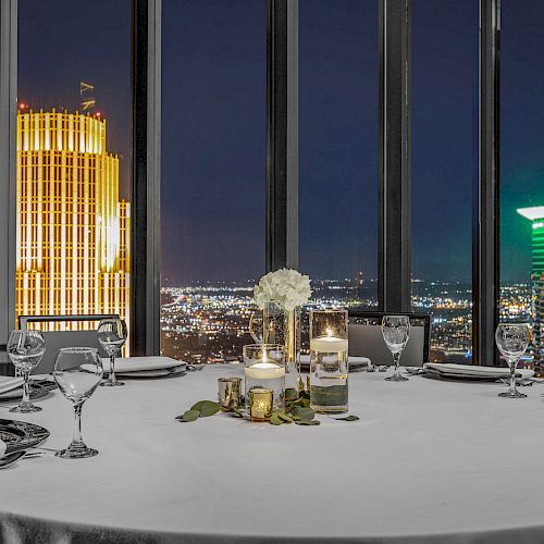 A dining table setup with white cloth, candles, glasses, and napkins in a room with a city night view through tall windows.