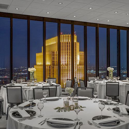 A beautifully set dining room with round tables overlooks a brightly lit skyscraper at night through large windows, offering a stunning city view.