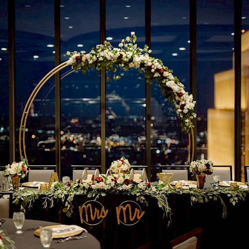 A beautifully decorated wedding table with floral arrangements, "Mr. & Mrs." signs, and a night cityscape view through large windows at the background.