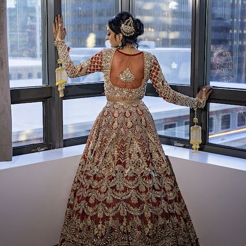 A person in a traditional embroidered outfit stands by a window, looking outside in an urban setting with tall buildings visible.