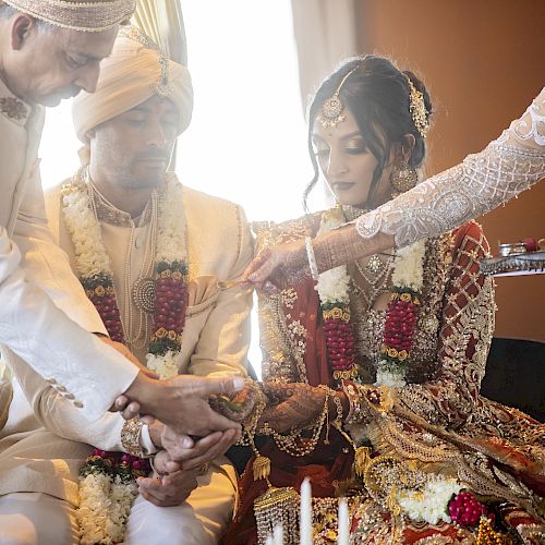 A traditional wedding ceremony where a couple, adorned in cultural attire, participates in ritualistic hand gestures with two individuals assisting them.