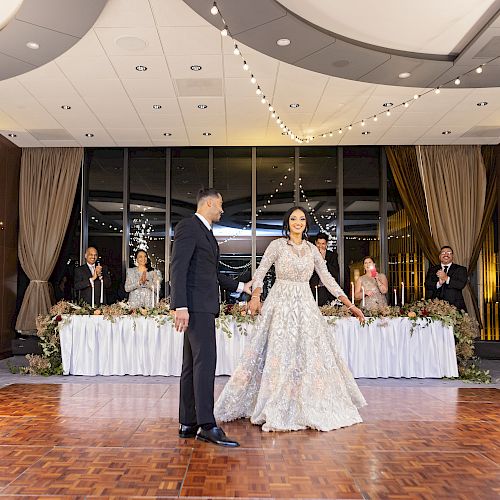 A couple, possibly newlyweds, are on a dance floor with sparklers around them. Guests seated at a table in the background are clapping.