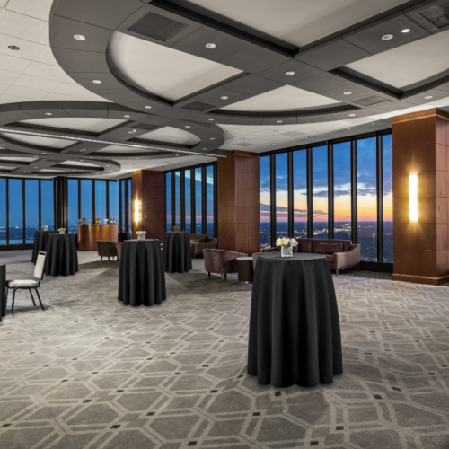 This image shows an elegant, empty event space with tall windows, cocktail tables with black tablecloths, and chairs, offering a stunning city view at sunset.