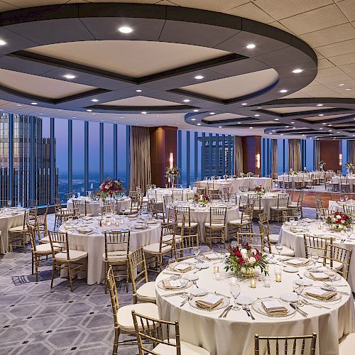 The image shows an elegant banquet hall with round tables set for an event, featuring floral centerpieces and city views through large windows.