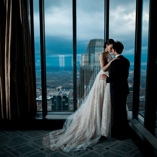 A couple in formal attire embraces near a tall window with a cityscape and cloudy sky in the background, creating a romantic atmosphere.