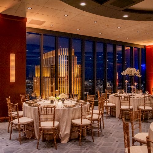 The image shows an elegant banquet hall set for an event, with round tables, chiavari chairs, and a city view through large windows at night.