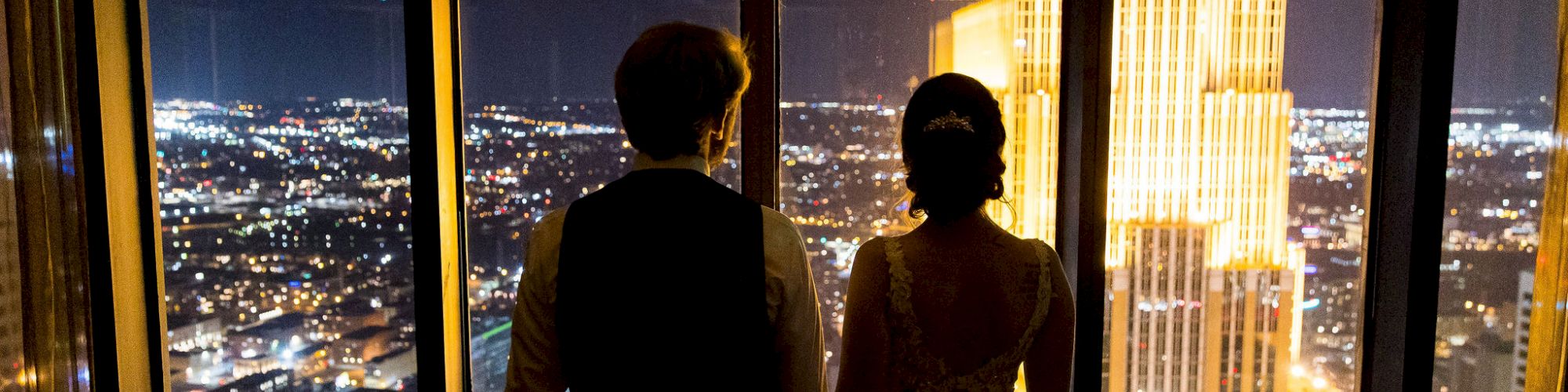 Two people are standing by a window at night, holding hands, overlooking a brightly lit city skyline.