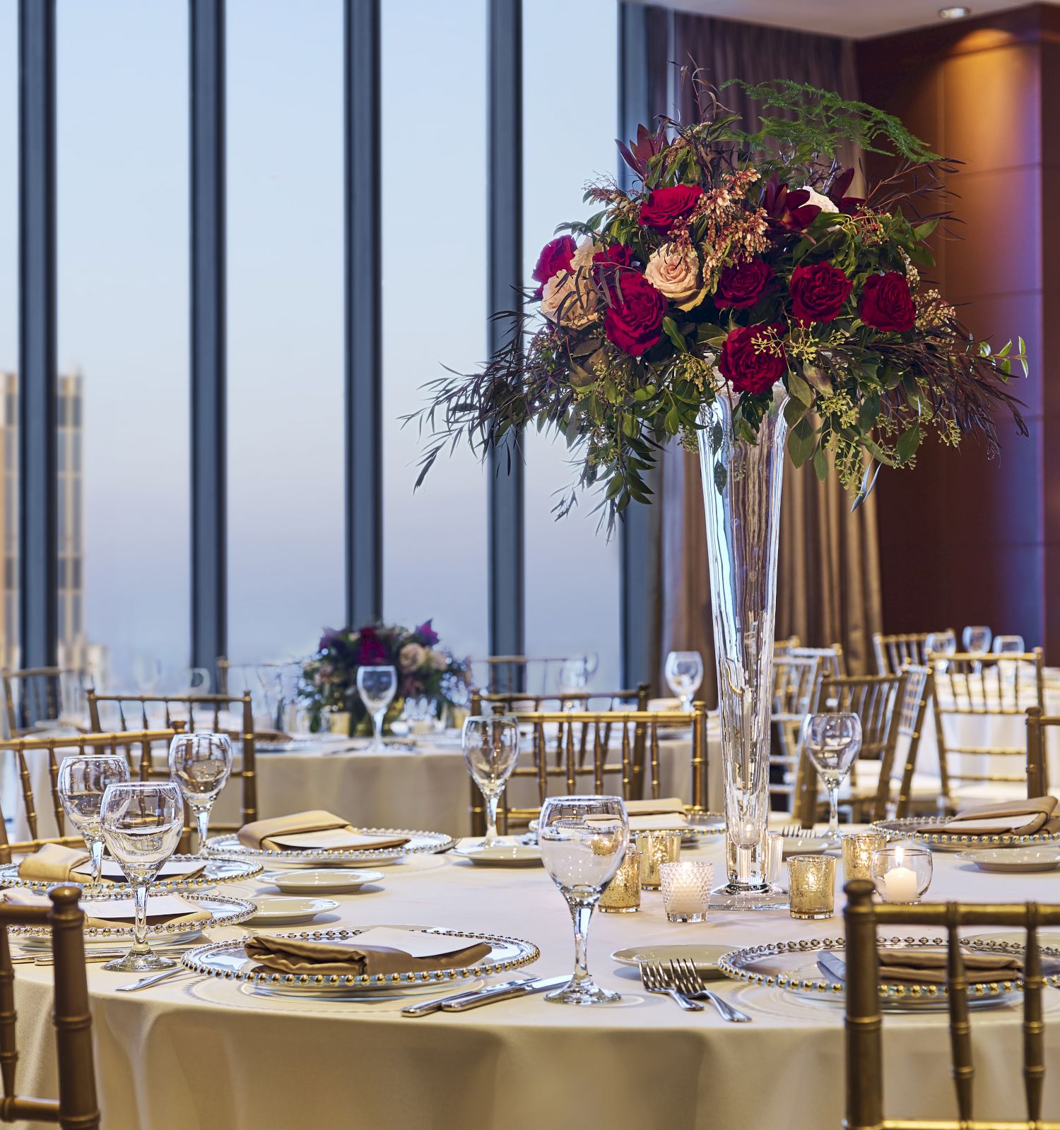 A beautifully set dining room with elegant tableware, floral centerpieces, and a view of tall buildings through large windows at sunset.