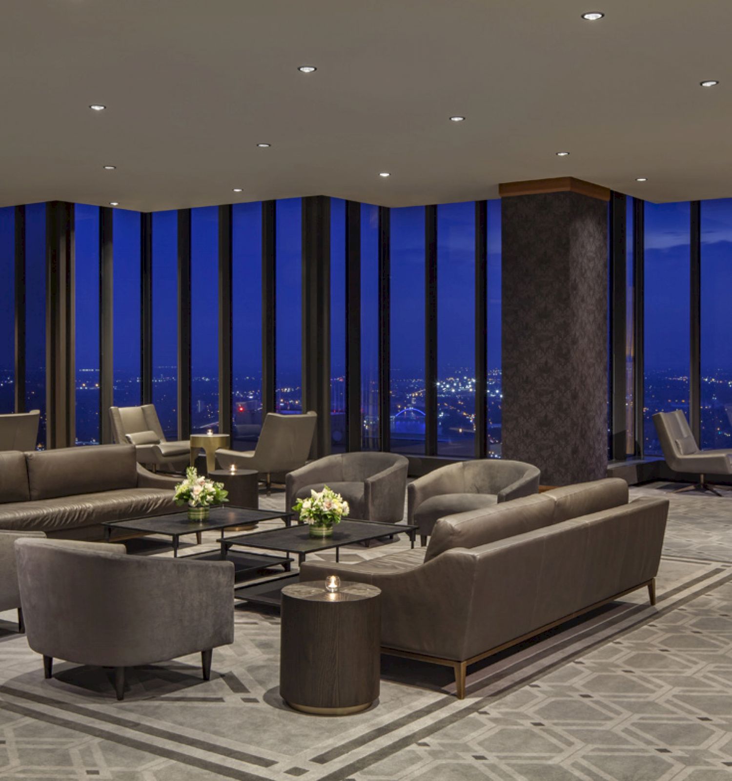 The image shows an elegantly furnished lounge area with modern sofas and chairs, large windows revealing a nighttime cityscape.