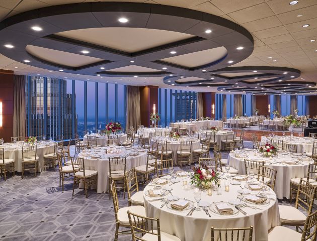 A decorated banquet hall with round tables, chairs, floral centerpieces, and elegant lighting, set up for a formal event with a city view.