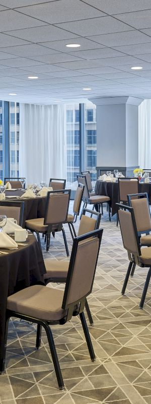 A banquet hall is set up with round tables, each adorned with folded napkins, glassware, and centerpieces. Large windows provide natural light.