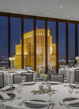 A modern dining room with round tables set for a formal event, featuring large windows with a city view and illuminated building outside at night.