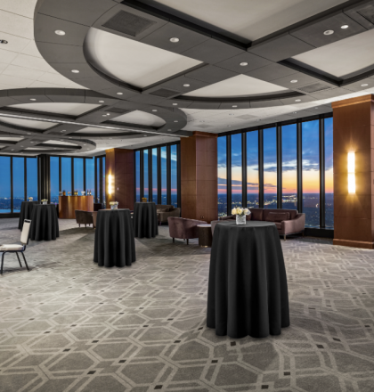 A spacious, modern event room with large windows providing a view of the sunset, filled with high tables and chairs, featuring elegant decor.