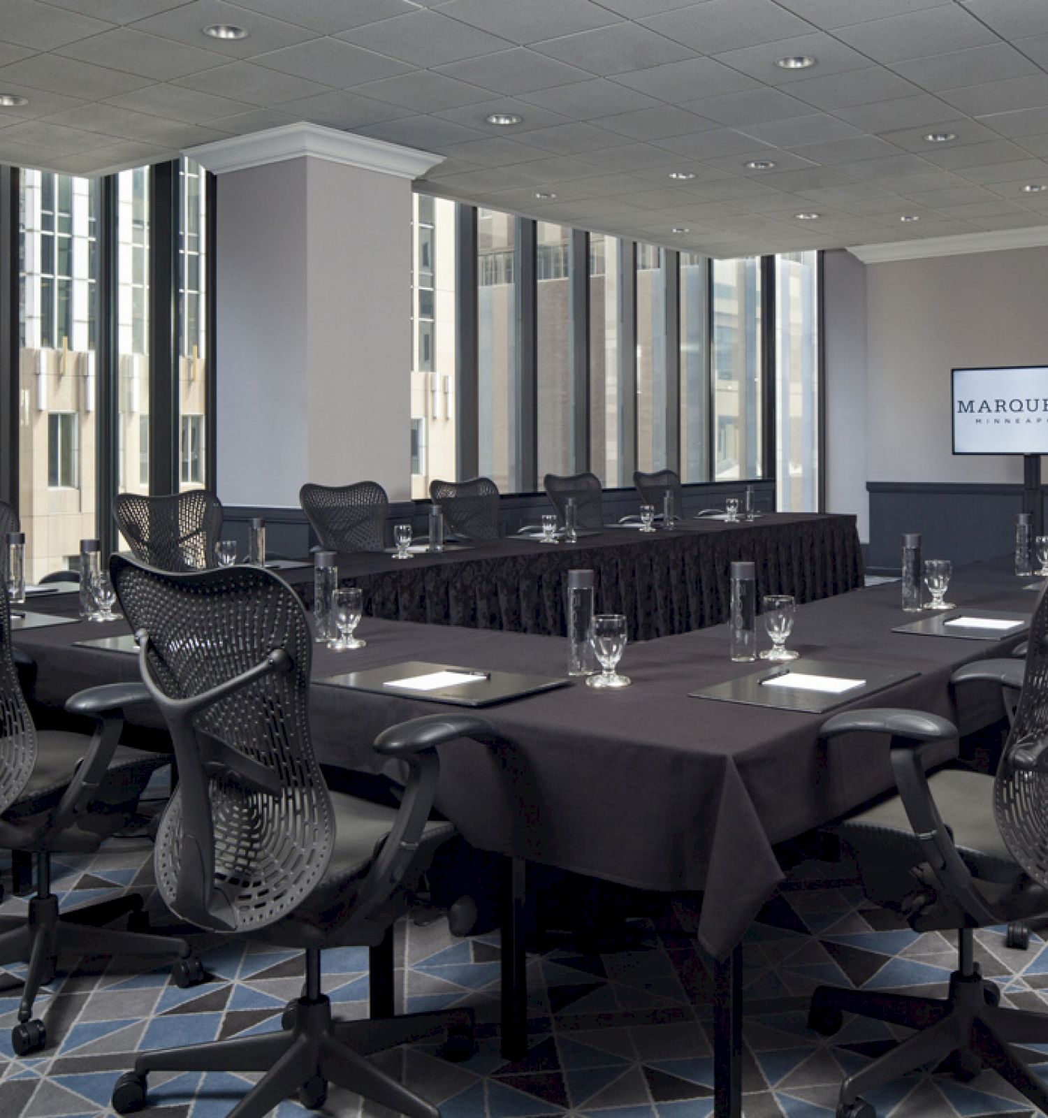 A modern conference room with large windows, black chairs, a long table with black tablecloths, glassware, and a screen displaying "MARQUETTE."