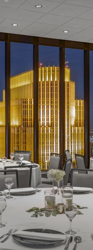 A formal dining room with round tables, elegant place settings, and large windows offering a night view of a brilliantly lit building.