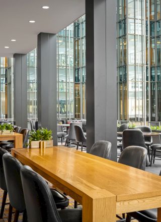 The image shows a modern indoor dining area with wooden tables and black chairs, large windows, and greenery, creating a comfortable and stylish ambiance.