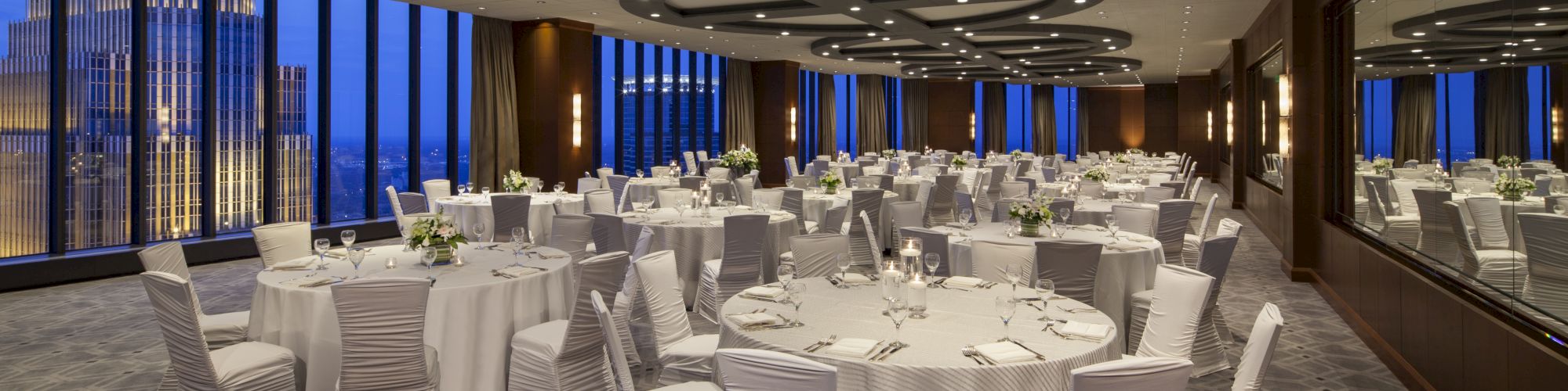 The image shows an elegantly decorated banquet hall with round tables, white chairs, and large windows offering a cityscape view at night.