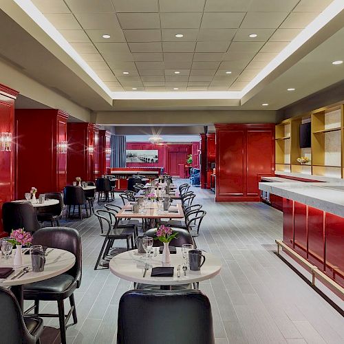 A modern restaurant with red decor and a long bar counter, featuring round tables with flowers and chairs neatly arranged in the seating area.