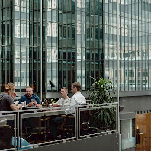 Four people are sitting around a table in a modern office building with glass windows, engaged in a discussion with notebooks and devices.