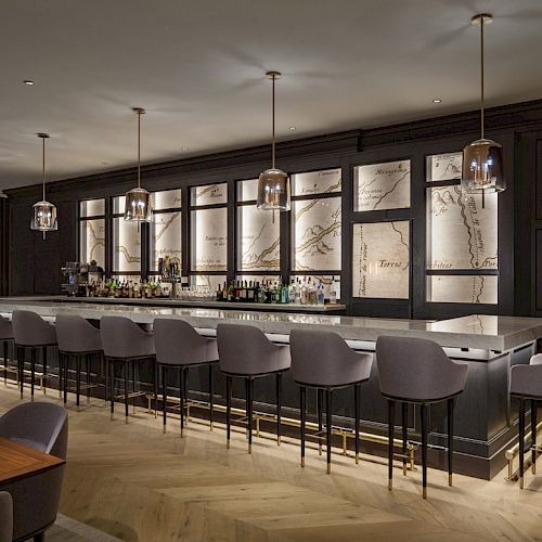 The image shows a modern bar with a sleek counter, high chairs, pendant lights, and a backdrop featuring illuminated panels.