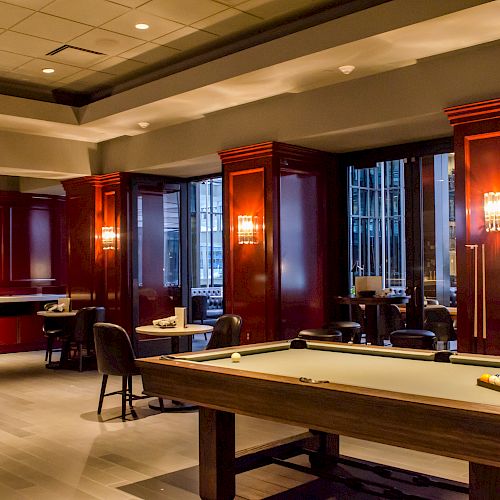 The image shows a stylish lounge with a pool table, dark wooden panels, classic furniture, and cityscape views through the windows.