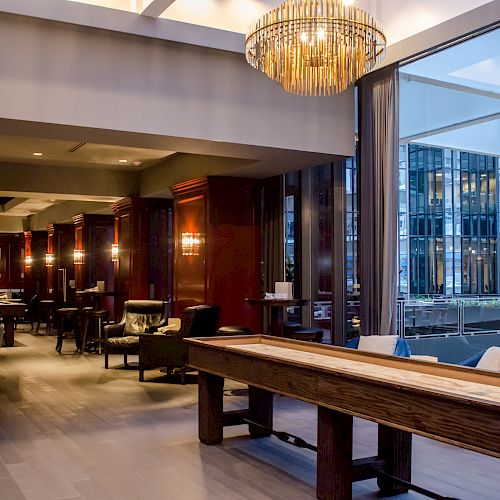 An indoor lounge area with elegant lighting, armchairs, tables, and a shuffleboard table; a large window offers a view of an adjacent building.