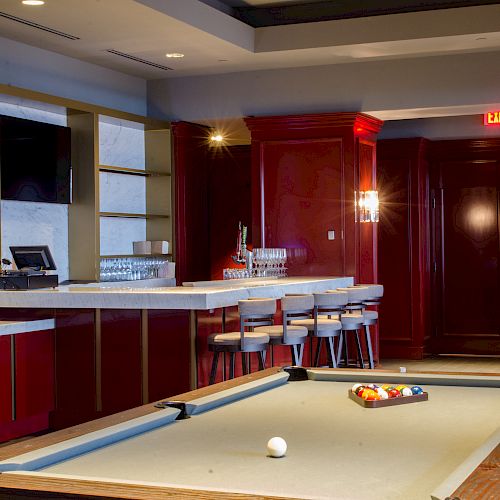 The image shows a bar area with a pool table in the foreground, and bar stools lined up in front of a counter.