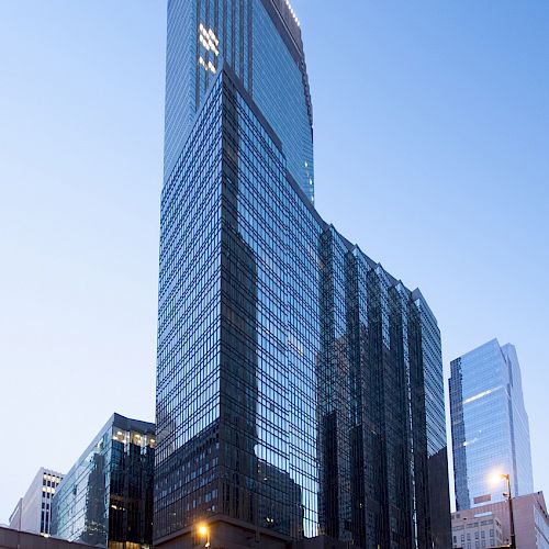 A tall, modern glass building stands prominently against a clear sky, with other buildings and streetlights visible in the foreground.