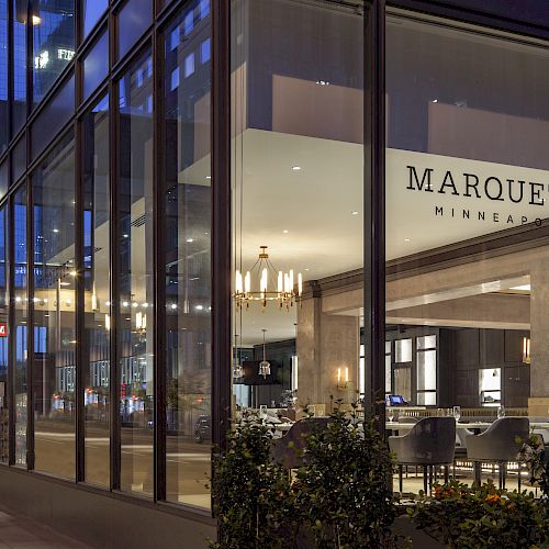 The image shows the exterior of Marquette Hotel in Minneapolis at night, featuring its well-lit entrance and modern glass facade.