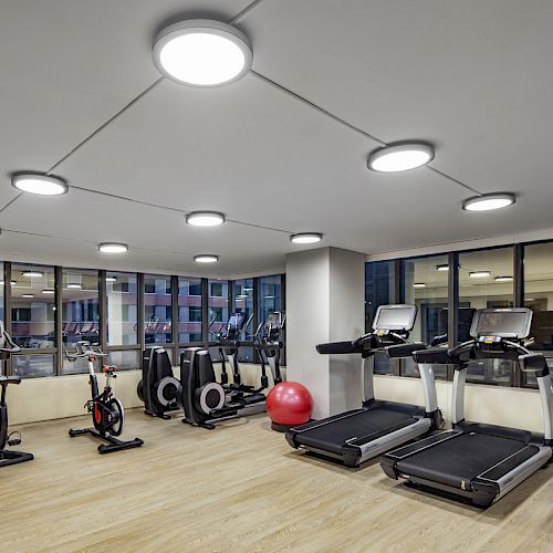 This image shows a gym with treadmills, exercise bikes, rowing machines, and a red exercise ball, all well-lit with ceiling lights.