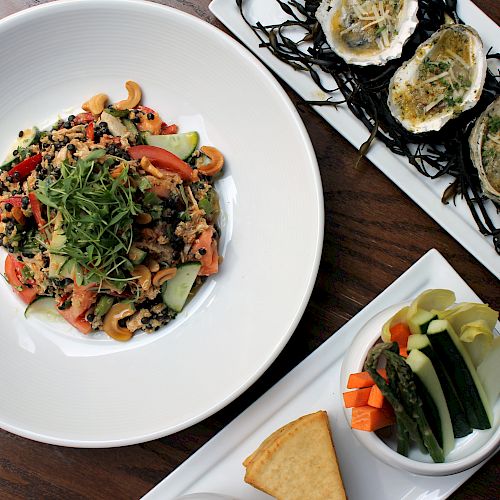 The image shows a table setting with a bowl of mixed salad, a plate of oysters, and a small dish of assorted pickles with a piece of bread.