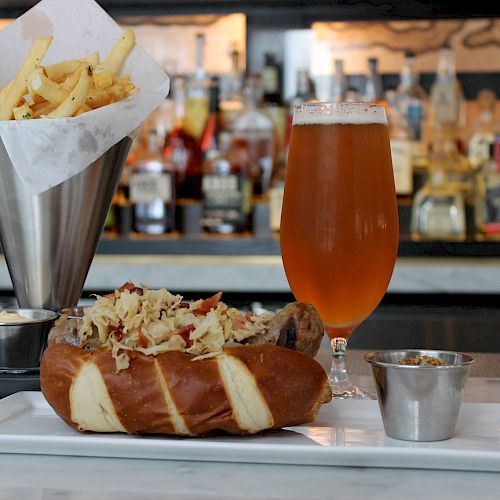 A plate with a hot dog topped with sauerkraut, a side of fries in a metal cup, small condiment containers, and a glass of beer on a bar counter.