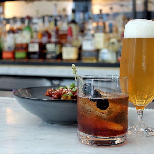 The image shows a bar scene with a glass of beer, a cocktail, and a bowl of food on a marble counter, with various bottles in the background.