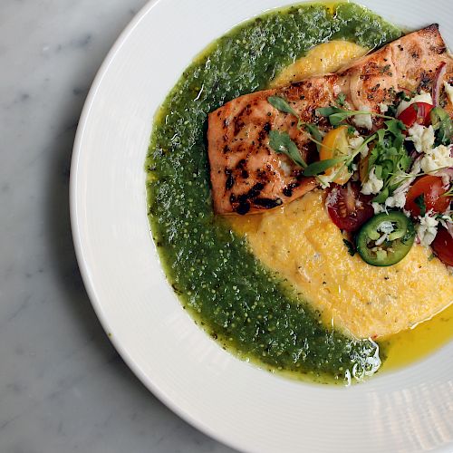 A grilled salmon fillet is served with creamy polenta and a fresh vegetable garnish, all surrounded by a vibrant green sauce.