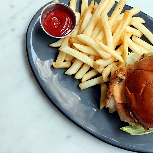 A burger with lettuce in a bun, served with a side of French fries and a small cup of ketchup on a dark plate.