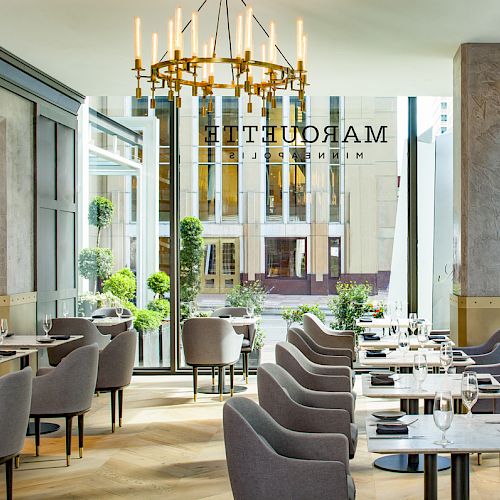 This image shows a modern, elegant restaurant with gray chairs and tables set for service. A chandelier hangs from the ceiling and there are large windows.