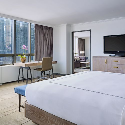 The image shows a modern hotel room with a large bed, a desk by the window, a TV on a dresser, and a separate seating area, offering city views.