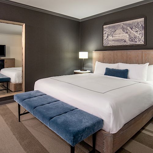 A modern hotel room with a large bed, blue bench, wall mirror, TV, lamps, wooden furniture, and a framed picture above the bed, well-lit and tidy.