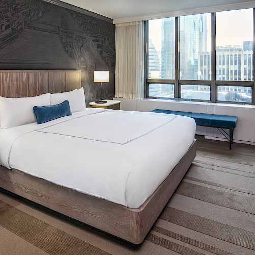 A modern hotel room with a large bed, two bedside tables, lamps, a TV on a dresser, and a window with city views.