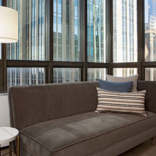 A modern living area with a gray sofa, decorative pillows, a floor lamp, and a small table. Large windows reveal a view of city skyscrapers.