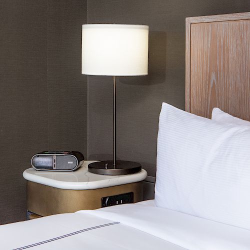 The image shows a bedside table with a lamp, an alarm clock, and part of a bed with white pillows and sheets against a wooden headboard.