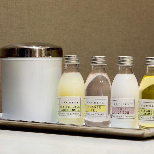 The image shows a tray with a white container and four small bottles of hotel toiletries, including conditioner, shower gel, body lotion, and shampoo.